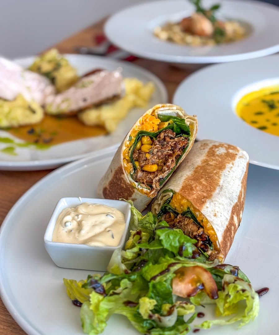 Grilovaný wrap s trhaným masem, kukuřicí a špenátem, se salátem a dipem; v pozadí další jídla a polévka